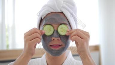 脸上戴着灰色面具的亚洲女人正在把几片黄瓜放在眼睛上，微笑着看着相机。水疗美容程序在家里为女孩。护肤、保湿及保湿概念.
